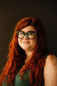 "A portrait of Hannah Wise, smiling slightly, wearing dark rimmed glasses, a green sleeveless top and long wavy red hair in front of a dark background."