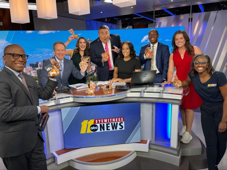 The ABC11 team stands and poses for a photo by the newsroom station desk.