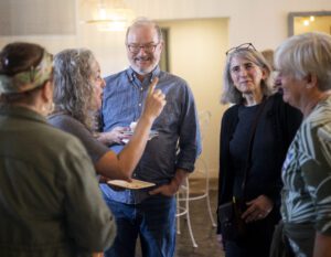 A house party showing five attendees wearing causal clothing and smiling, in conversation, with one raising her arm and gesticulating. 