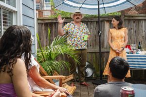 Hanna stands to the right in a small courtyard, wearing a melon-colored sun dress and next to a house party host who is wearing a patterned shirt and gesticulating. House party attendees sit in the foreground.