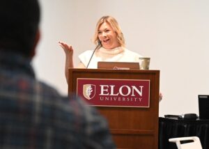 Shannan Bowen stands behind a podium with Elon University on the front. She is smiling, gesticulating and wearing cream top with a blond bob. 