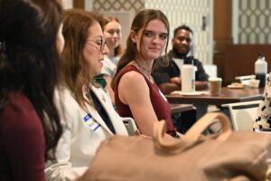 An image of RFA corps members sitting in front of tables listening to one another speak. In the middle is CityView's Morgan Casey, looking toward the camera and wearing a sleeveless burgundy top and long brown hair. 