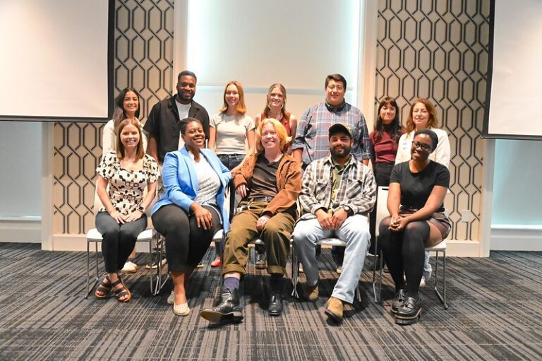 A photo of the 12 RFA corps reporters for North Carolina, five sitting in a row of chairs and seven standing behind them. They're pictured in a conference room at Elon University, and all are smiling.