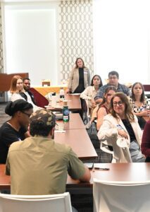 Eight RFA corps members sit around tables in a conference room, looking at one person who is talking. In the far back, program lead Sarah Day Owen Wiskirchen is standing in front of large windows. 