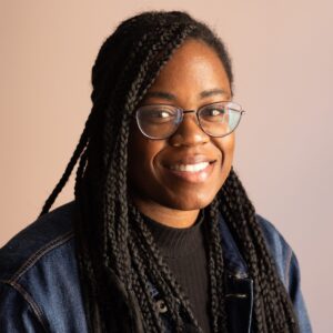 A portrait of P.R. Lockhart, smiling, wearing glasses and long dark brown braids. She's wearing a blue denim jacket and a black top.