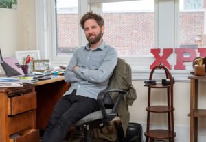 Thomas Calder leans back slightly in a desk chair, with arms folded. He's in the Mountain Express office, in front of big windows, wearing a light blue shirt, tousled brown hair and a beard. On the right, you can see large red letters sitting on the window sill spelling out Xpress.