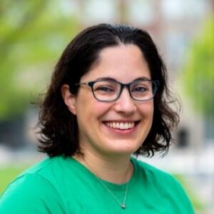 A portrait of Melody Joy Kramer pictured outside, smiling and wearing a bright green t-shirt, brown bob and glasses. 