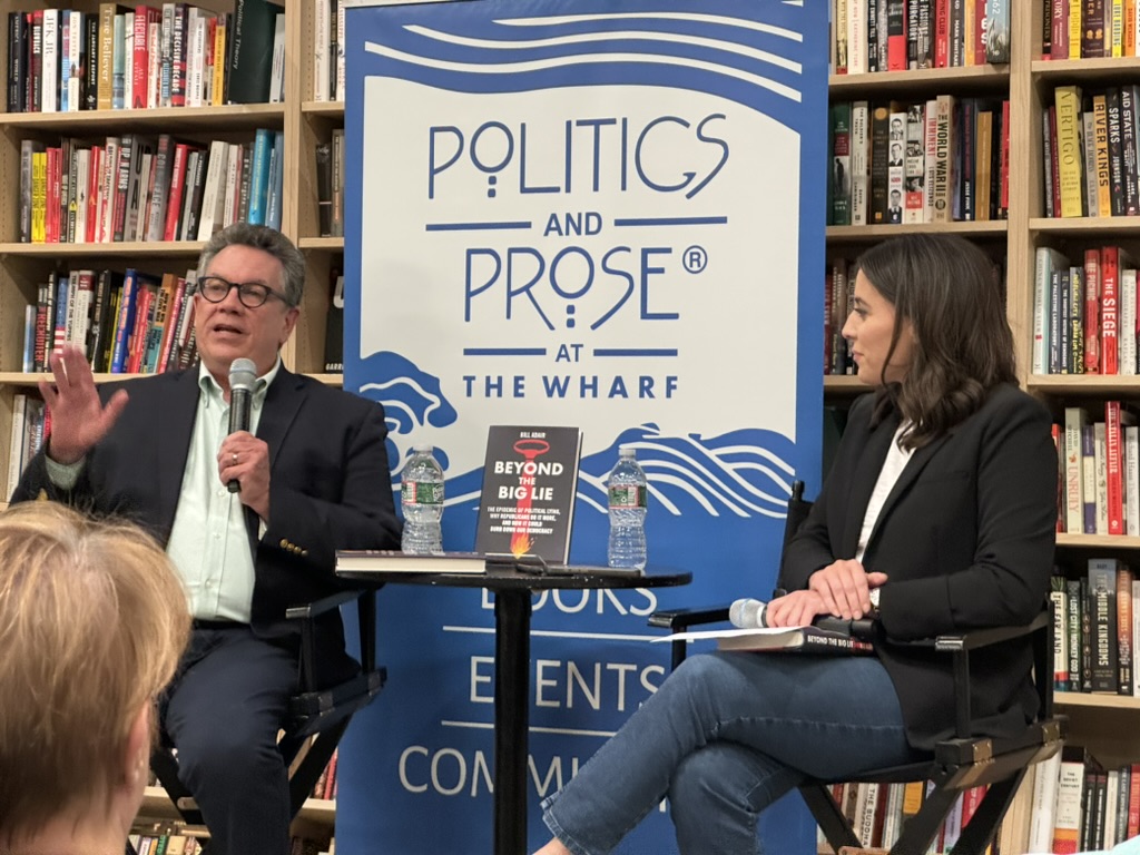 Bill Adair, seated left and wearing a suit while holding a microphone gesticulates to the audience. Next to him is Hallie Jackson wearing a dark blazer and blue jeans. Behind them is a wall of books and a blue and white Politics & Prose vertical banner.