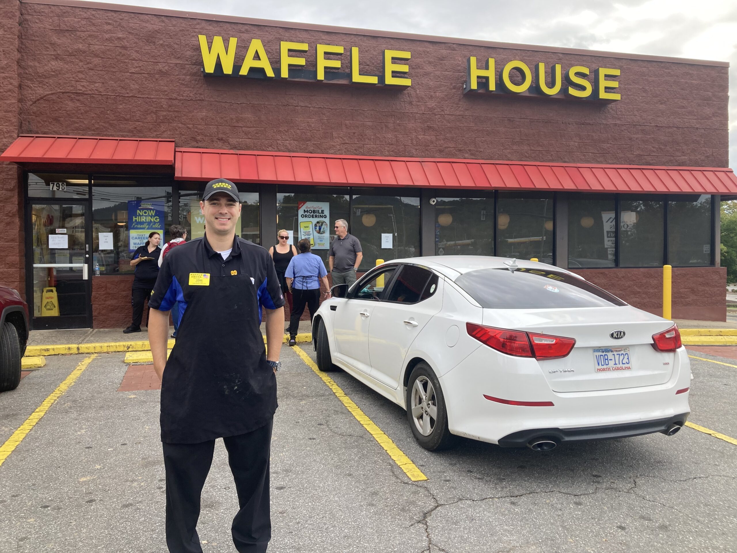 Logan Craig, wearing a Waffle House navy blue uniform and baseball hat stands in the parking lot in from of the restaurant. A white car and a few people are pictured to his right.