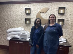 Wearing navy blue uniforms, Andrea Bouknight, left, and Heather Mallette, right, stand behind the front desk of Comfort Suites. They are smiling and there's a stack of white blankets and towels on a cabinet behind them.