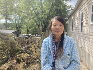 Meiling Dai stands next to her beige mobile home wearing a light blue robe over her clothes. Her long dark hair is in a braid. Behind her is a pile of tree debris from Tropical Storm Helene.