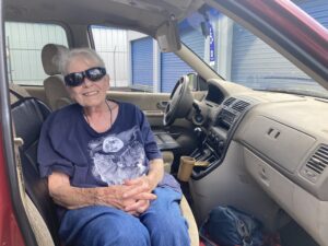 Barbara Biesterfeld sits in the passenger seat of a minivan, wearing a blue tee with a wolf and moon graphic, dark sunglasses and gray hair that's pulled back. She is smiling and folding her hands in her lap.