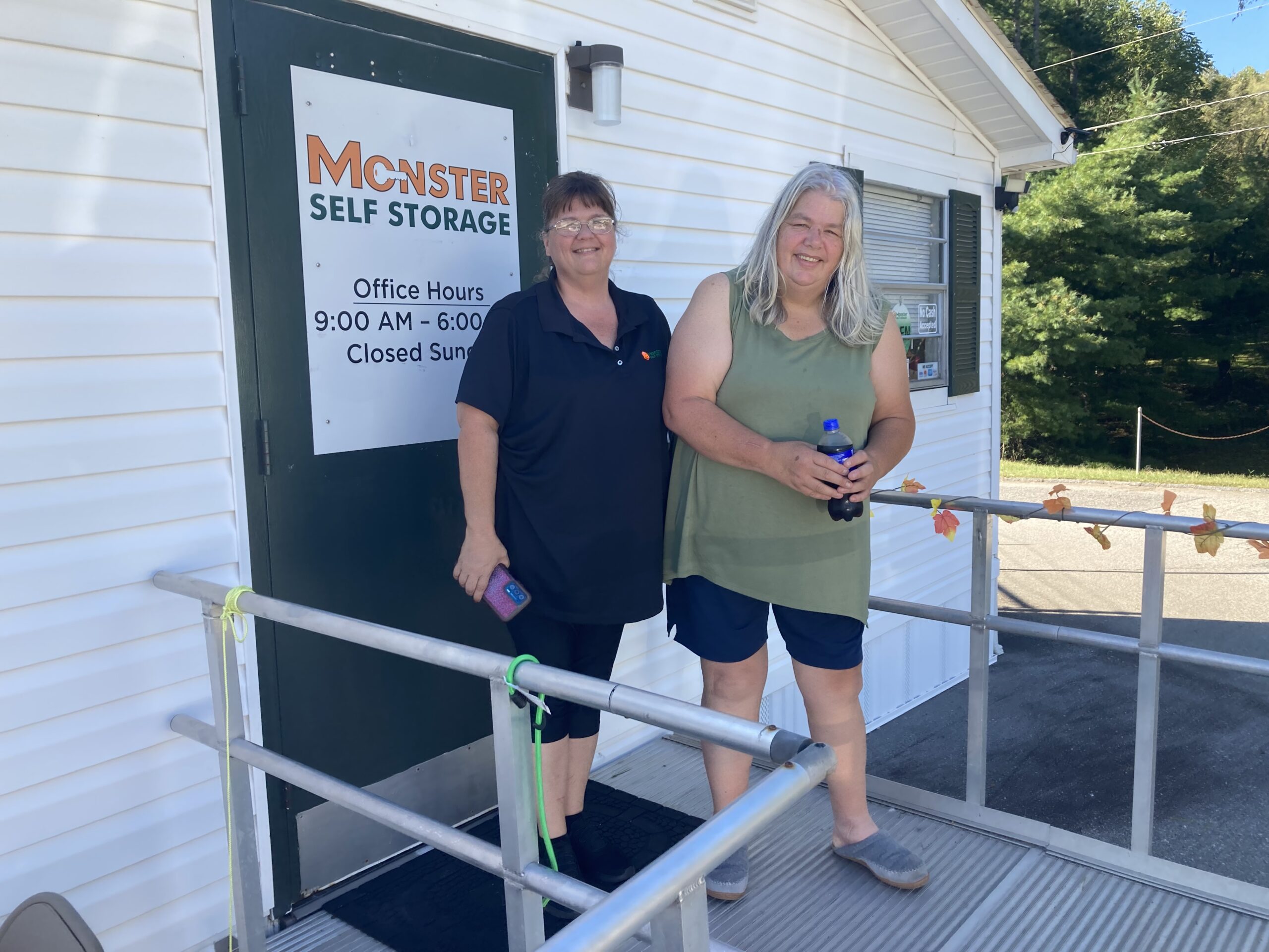 Keri Scott, wearing a dark polo and capri leggings and Michelle Fisher, wearing a moss green tank and dark shorts, smile and stand on the ramp in front of the small Monster Storage and Camelot Mobile Home office.