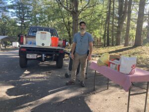 Camelot resident and facilities volunteer Arnold wears beige cargo pants, a blue tee, a long white beard and baseball hat, stands in front of a pick up truck with a large container of potable water and next to a table and grill set up to feed the community. In the background are white pines.