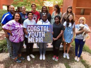 A dozen people, including adults and youth stand outside by a brick building with some trees and plants in the background. The group is diverse in race/ethnicity. One youth is holding a small white dog. A youth in the middle is holding a large handmade sign reading "Coastal Youth Media."