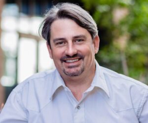 A portrait of Pate McMichael wearing a light Oxford shirt, light brown hair and a mustache and goatee, smiling and pictured outdoors.