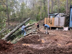 Gerard Albert III is pictured wearing a blue shirt and light pants, leaning over and photographing two people trying to move a pile of long tree trunks that have been partially chainsawed. They are all surrounded by mud and a damaged structure is seen in the back right.