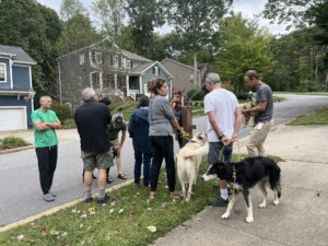 About 8 people stand outside on the sidewalk and street of a neighborhood, listening to a single radio set up on a post. It's cloudy, the group is wearing t-shirts, shorts and sweat pants, and there are two larger dogs pictured, one light beige, the other mostly black with some white on the legs and face.