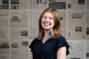 A portrait of Caroline Wills smiling and wearing a dark short sleeved top. She is standing in front of a wall of DTH newspapers. 