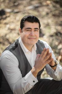 A portrait of Diego Barahona wearing a white button down shirt, gray vest, short dark hair and holding his hands together in an upside down V shape. 