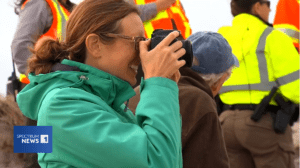 A side shot of Saunders, wearing a green rain coast, photographing something in the distance. A person in a bright yellow safety jacket can be seen in the background.