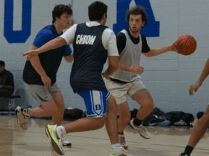 Three student journalists wear shorts and t-shirts while playing a game of basketball. One player is dibbling and another has a 