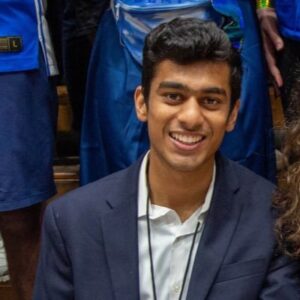 A portrait of Ranjan Jindal smiling and wearing a dark blue blazer and light top. He is sitting and appears to be at an athletic event covering a game.