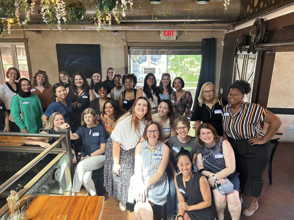 A group of about 30 people pose for a picture at the first NC Women in Journalism meet up. They are in a restaurant with a window in the back.