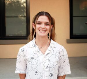 Morgan Casey stands outside a beige colored building. She is smiling, with long brown hair and wearing a white collared top with a pattern on it.